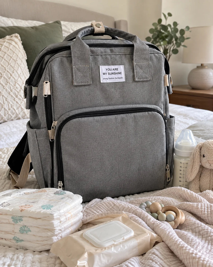 The grey backpack on a bed with nappies, a bottle and a soft toy